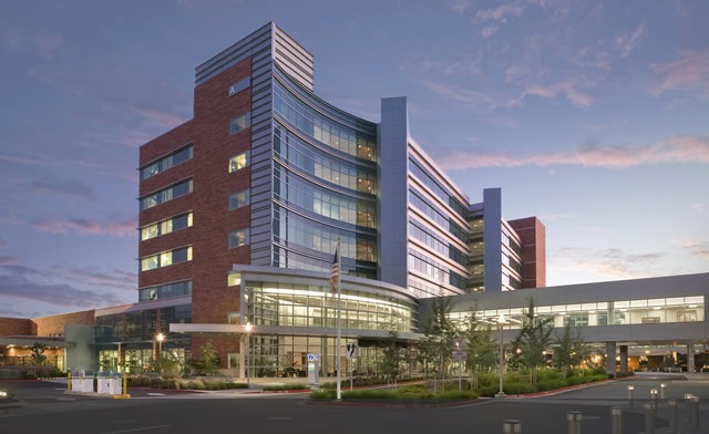 A modern hospital building with multiple stories, large glass windows, and a curved entrance, seen at dusk with lights on inside and a partly cloudy sky in the background.