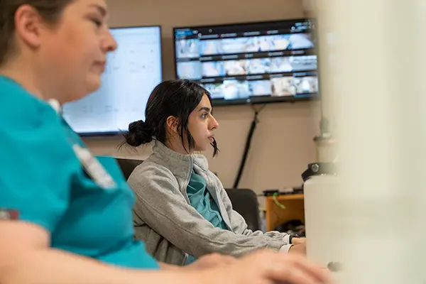 Two women in medical scrubs sit at desks and work on computers in an office with surveillance monitors displayed on the wall behind them.