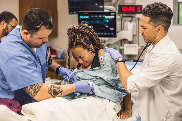 A woman in labor is supported by two medical professionals, one in blue scrubs and one in a white coat, as she sits on a hospital bed during childbirth. Medical monitors are visible in the background.