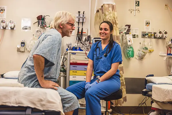 A nurse in blue scrubs smiles while talking to an older male patient sitting on a hospital bed in a medical examination room with various medical equipment in the background.