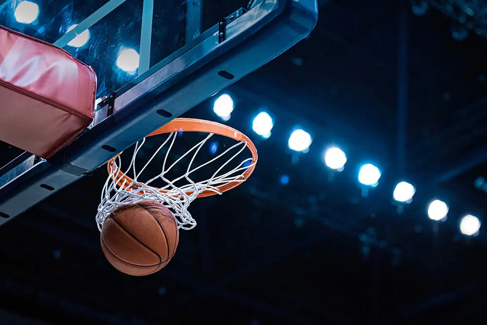 A basketball passes through the hoop and net from below, with bright arena lights shining in the background.