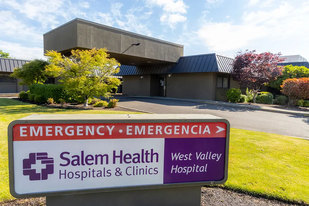 A sign reading Emergency • Emergencia, Salem Health Hospitals & Clinics, West Valley Hospital stands in front of a modern hospital building surrounded by trees and grass on a sunny day.