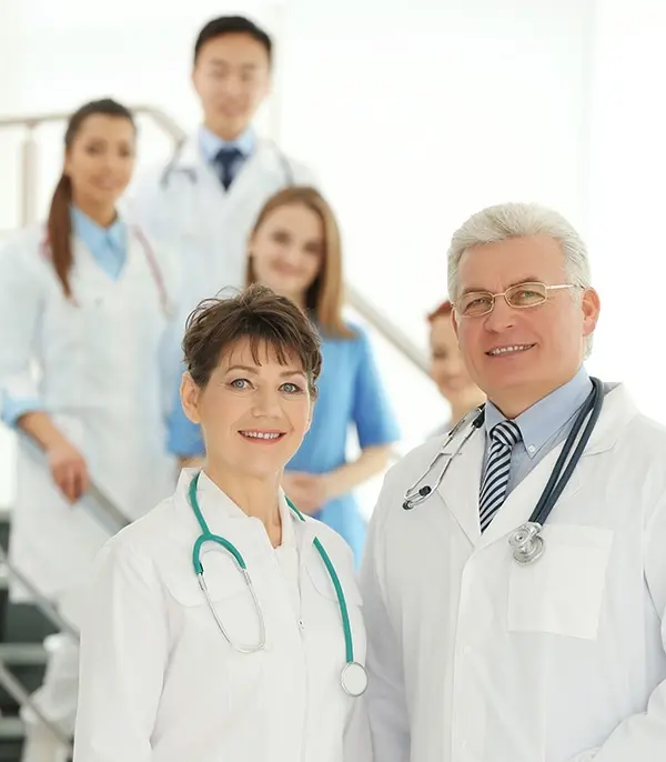 Two doctors wearing white coats and stethoscopes stand in front, smiling, with three other medical professionals and a nurse standing in the background on a staircase. All appear friendly and professional.